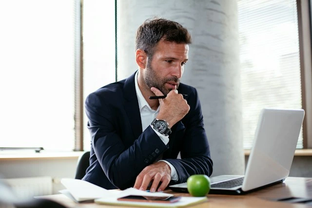 Un hombre de negocios con traje pensativo mirando la pantalla de su computadora portátil en una oficina brillante.
